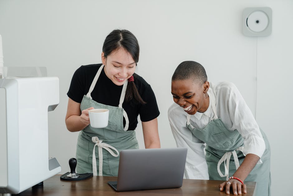 Happy young Asian and African American female coffee house coworkers in aprons laughing while browsing Internet on modern laptop during break