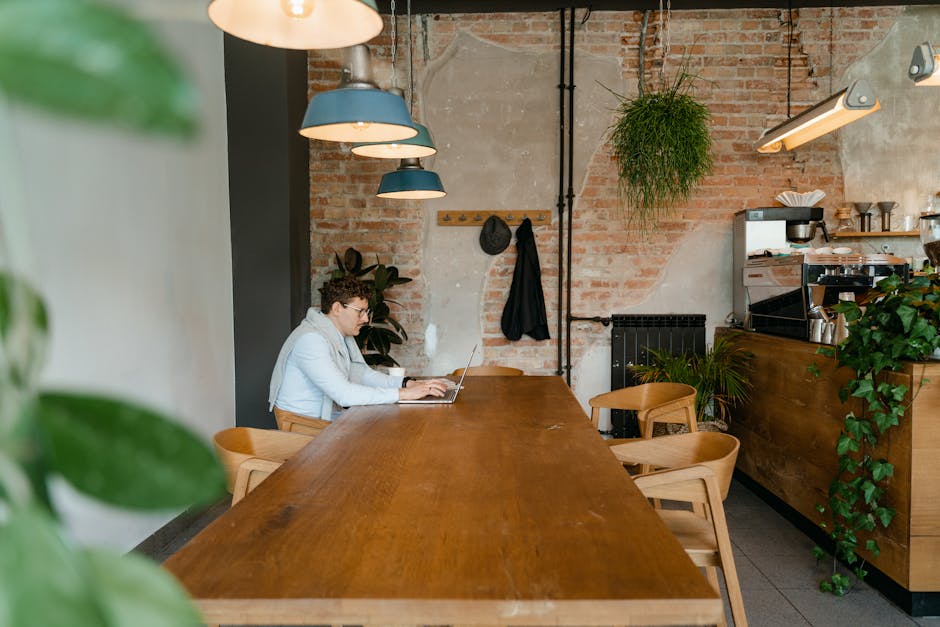 A man working on a laptop in a cozy cafe with greenery and wooden decor.