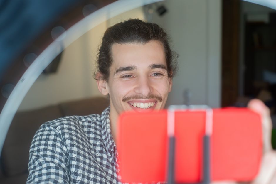 Happy man taking selfie for vlogging using smartphone and ring light setup indoors.