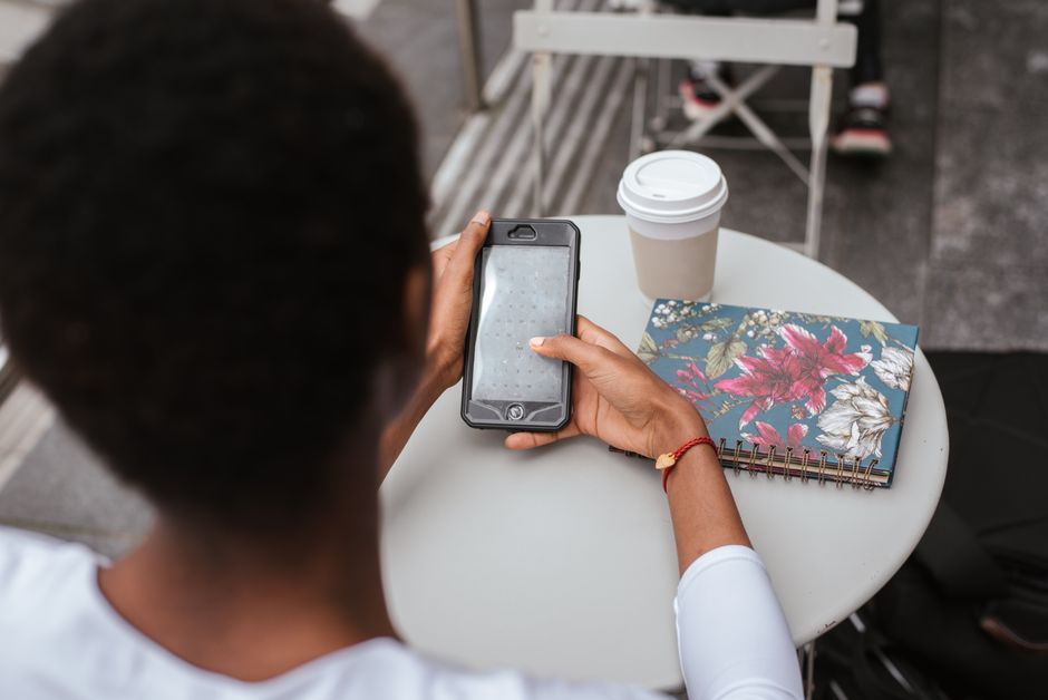 High angle back view of faceless African American lady sitting at table in cafeteria and checking notification on smartphone near notebook and cup of drink