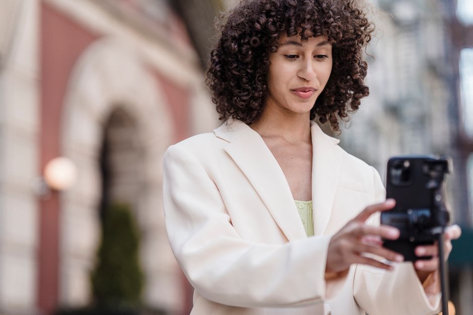 Young ethnic female vlogger with curly hair in jacket preparing cellphone for video record in town