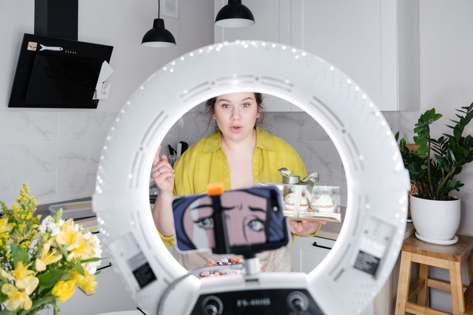 Young female blogger standing in kitchen with box of sweets in front of smartphone placed on tripod with ring lamp while recording vlog