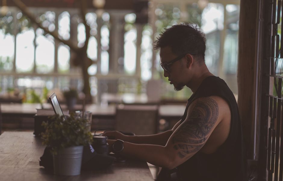 Side view of a tattooed man with eyeglasses working on a laptop in a cafe.