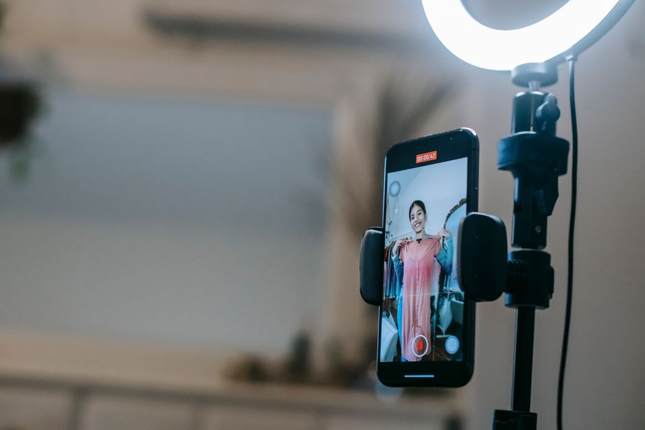 Anonymous female blogger standing in room and shooting vlog on smartphone with ring lamp while demonstrating pink dress in hands