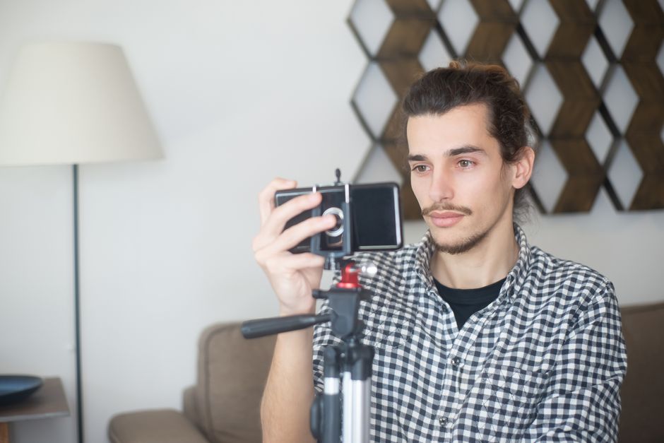 Young man with plaid shirt using a smartphone and tripod to create content indoors in Portugal.